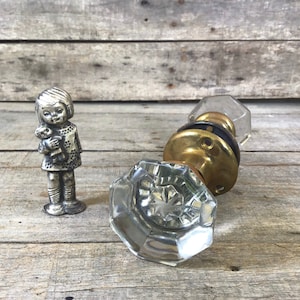 May include: A silver figurine of a girl holding a doll stands next to two vintage glass and brass doorknobs. The doorknobs have a faceted crystal design. The items are arranged on a weathered wooden surface.