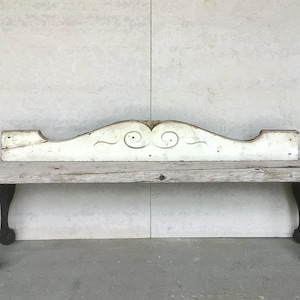 May include: A long, horizontal wooden architectural element with a distressed white painted finish. The piece features a central arched top with carved scrollwork details and symmetrical curved lines, showing visible signs of wear and aging.