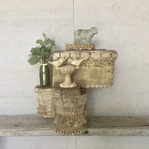 May include: A rustic, weathered wooden shelf with decorative elements. It features a small sheep statue, a bird bath with two bird figurines, and a green glass bottle with faux greenery. The shelf has a distressed, aged appearance.