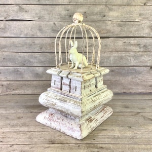 May include: A decorative white and distressed-finish birdcage-style sculpture with a small white rabbit figurine inside. The sculpture sits atop a multi-tiered, aged-look pedestal. The cage has a round top with a small ball finial.