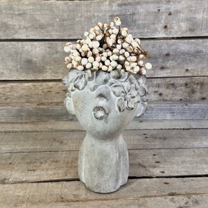 May include: A gray, decorative head-shaped planter with a textured finish, filled with white berries. The planter has detailed facial features and floral accents around the head. The background is a rustic wooden surface.