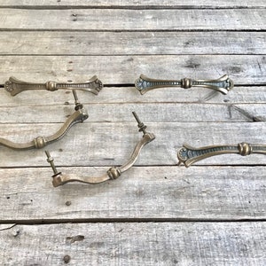 May include: Antique brass drawer pulls and handles. The hardware includes two straight pulls and three curved pulls. The pulls have decorative detailing and are mounted with screws and nuts. The background is weathered wood.