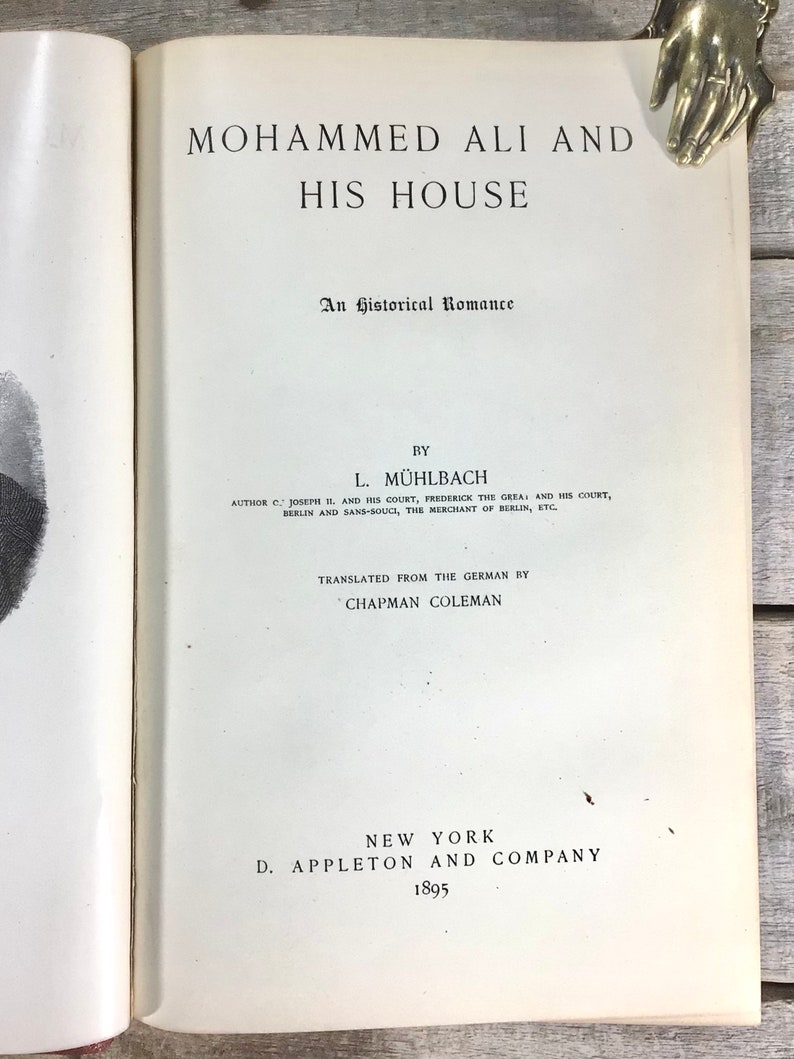 Puede incluir: Libro abierto titulado "Mohammed Ali and His House" con texto negro. El libro es una novela hist&oacute;rica de L. M&uuml;hlbach, traducida del alem&aacute;n por Chapman Coleman, publicada en 1895 por D. Appleton and Company, Nueva York.
