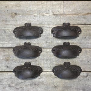 May include: Six antique-style cast iron drawer pulls with a dark brown finish. Each pull has a decorative leaf design and two screw holes for mounting. The pulls are arranged on a weathered wooden surface.