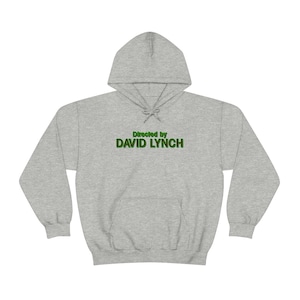 Puede incluir: Una sudadera con capucha gris con el texto "Directed by DAVID LYNCH" en letras verdes con contorno.