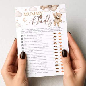 Op de afbeelding: Een babyshower-spelkaart met de tekst "Mummy or Daddy" en een teddybeer-illustratie. De kaart bevat vragen over de aanstaande ouders, met snor- en lipafbeeldingen om te beantwoorden. De kaart wordt door een persoon vastgehouden.