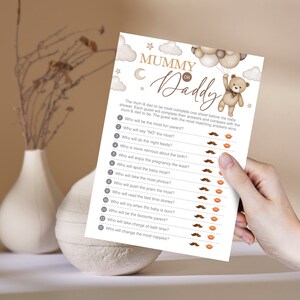 Może przedstawiać: Drukowana karta gry na baby shower z tytułem "Mama czy Tata?" Karta przedstawia brązowego pluszowego misia trzymającego balony i listę 13 pytań dotyczących przyszłych rodzic&oacute;w. Obok każdego pytania znajduje się ikona wąs&oacute;w lub ust, aby goście mogli zaznaczyć swoje odpowiedzi.