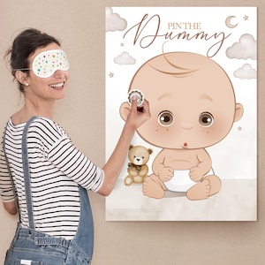 Op de afbeelding: Een babyshower-spel met een grote ge&iuml;llustreerde baby met een luier, een teddybeer en de tekst "Pin the Dummy". Een persoon met een slaapmasker probeert een sticker op de baby te plakken.