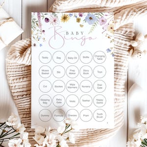 Op de afbeelding: Een Baby Bingo-spelkaart met bloemaccenten in pastelkleuren. De kaart heeft een raster van cirkels met babygerelateerde woorden zoals "Teddy", "Fles" en "Wandelwagen". Het woord "Baby Bingo" staat in een sierlijk lettertype bovenaan.