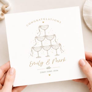 Könnte beinhalten: Eine quadratische weiße Grußkarte mit einer goldenen Strichzeichnung eines Champagnerglasturms. Der Text lautet CONGRATULATIONS, Emily & Mark, und 23RD JUNE 2026. Kleine goldene Herzen und Punkte sowie ein kleiner Blattzweig verzieren das Design.