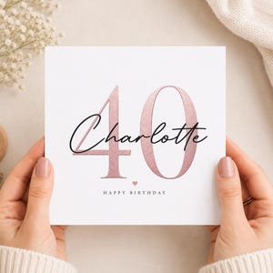 Könnte beinhalten: Eine quadratische weiße Grußkarte mit der großen Zahl 40 in roségoldener Metallic-Optik. Der Name Charlotte ist in schwarzer Schreibschrift in der Mitte platziert. Darunter befindet sich ein kleines roségoldenes Herz und der Text HAPPY BIRTHDAY.