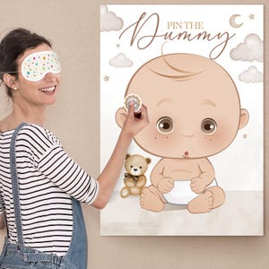 May include: A "Pin the Dummy" baby shower game poster featuring a cartoon baby with a teddy bear. The poster has a beige background with clouds, stars, and the text "Pin the Dummy". A person is shown wearing a sleep mask and attempting to pin a sticker.