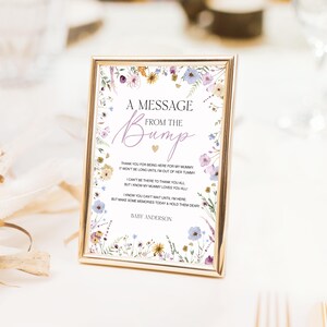 K&ouml;nnte beinhalten: Ein goldgerahmtes Schild mit dem Text "A Message From The Bump" in eleganter Schrift. Das Schild ist mit einem floralen Rand aus lila, gelben und blauen Blumen verziert. Der Text enth&auml;lt eine Nachricht vom Baby und den Namen "Baby Anderson".