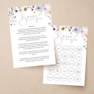 Op de afbeelding: Twee babybingo-spelkaarten met bloemaccenten in paars, geel en wit. E&eacute;n kaart heeft instructies en de andere heeft bingovakken met babygerelateerde woorden zoals "fles" en "speelgoed". De woorden "Baby Bingo" zijn in cursief geschreven.