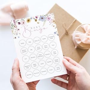 Op de afbeelding: Een Baby Bingo-kaart met bloemaccenten in paars, geel en blauw. De kaart heeft een raster van cirkels met babygerelateerde woorden zoals "Teddy", "Fles" en "Wandelwagen". De kaart wordt door een persoon vastgehouden.