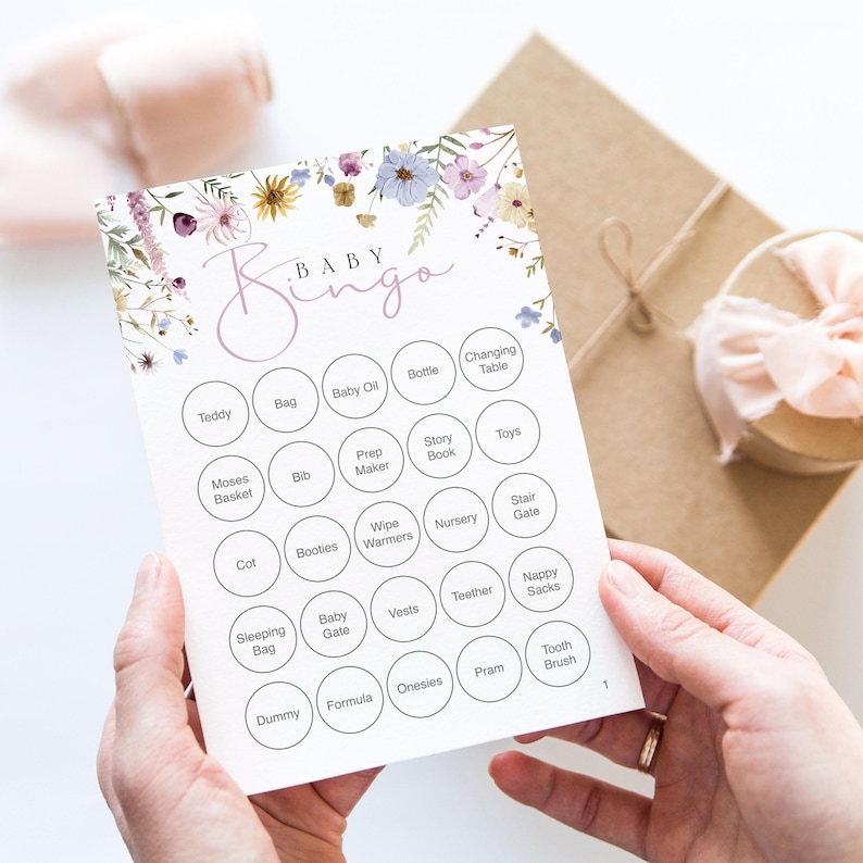 Op de afbeelding: Een Baby Bingo-kaart met bloemaccenten in paars, geel en blauw. De kaart heeft een raster van cirkels met babygerelateerde woorden zoals "Teddy", "Fles" en "Wandelwagen". De kaart wordt door een persoon vastgehouden.