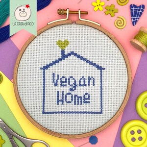 Puede incluir: Un bastidor de bordado de punto de cruz con las palabras "Vegan Home" bordadas en hilo azul. El diseño presenta un contorno de casa con un detalle en forma de corazón. El bastidor de madera está rodeado de suministros de costura, incluyendo botones e hilo.