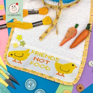 Puede incluir: Un babero blanco con un ribete floral amarillo y un diseño de punto de cruz de dos pollitos amarillos y el texto "FRIENDS NOT FOOD". La imagen también incluye hilo de bordar amarillo, botones decorativos y dos zanahorias naranjas.