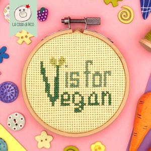 Puede incluir: Un bastidor de bordado de punto de cruz con las palabras "V is for Vegan" en hilo verde. El bordado está sobre una tela amarilla clara. El bastidor de madera está rodeado de materiales de manualidades coloridos sobre una superficie rosa.