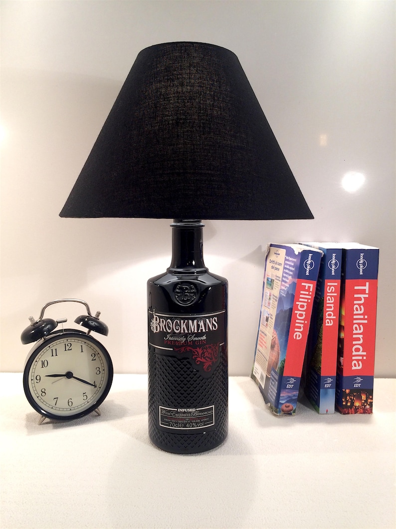 May include: A black lampshade with a black glass bottle base. The bottle is labeled "Brockmans Intensely Smooth Premium Gin" and has a red and gold design. The lamp is sitting on a white surface with a black alarm clock and three travel guide books.