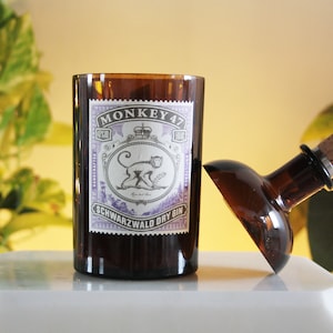 Può includere: Un bicchiere marrone con un'etichetta che recita "MONKEY 47 SCHWARZWALD DRY GIN" e una bottiglia con un tappo di sughero. L'etichetta presenta un'illustrazione di una scimmia. Il bicchiere e la bottiglia sono su una superficie bianca.