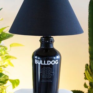 May include: A black lamp with a black fabric lampshade. The base of the lamp is a repurposed Bulldog Gin bottle with the brand name and description printed on the bottle in white.