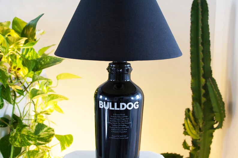 May include: A black lamp with a black fabric lampshade. The base of the lamp is a repurposed Bulldog Gin bottle with the brand name and description printed on the bottle in white.