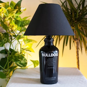 May include: A black lamp with a black lampshade sits on a white surface. The lamp base is a repurposed gin bottle with the label "Bulldog London Dry Gin".