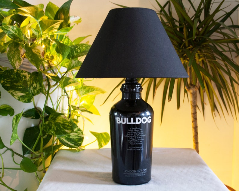 May include: A black lamp with a black lampshade sits on a white surface. The lamp base is a repurposed gin bottle with the label "Bulldog London Dry Gin".