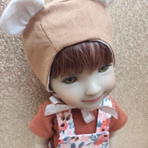 Peut inclure: Une poupée aux cheveux bruns portant un chapeau brun et blanc avec des oreilles d'ours et une robe à bretelles à imprimé floral.