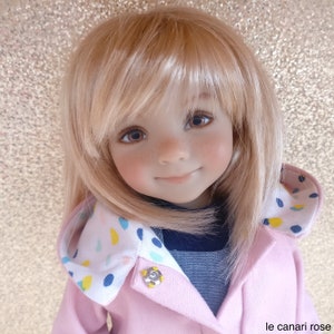 Puede incluir: Una muñeca con cabello rubio claro y ojos marrones, vestida con un abrigo rosa con capucha estampada. El abrigo tiene un cierre de botón a presión plateado. La muñeca tiene pecas y una sonrisa suave. El texto "le canari rose" está en la esquina inferior derecha.