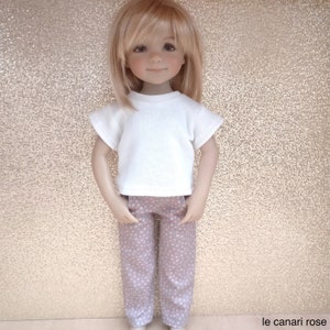 Peut inclure: Une poupée portant un t-shirt blanc et un pantalon gris à pois roses. La poupée a les cheveux blonds et les yeux bleus.