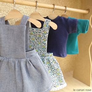 Peut inclure: Trois robes de poupée suspendues à des cintres en bois. La robe de gauche est une robe en jean bleu avec une salopette. La robe du milieu est une robe blanche à motif floral. La robe de droite est un t-shirt bleu et vert.