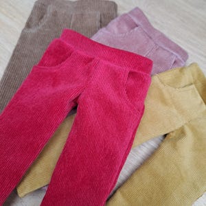 Peut inclure: Quatre paires de pantalons en velours côtelé de différentes couleurs : marron, rose, rouge et jaune moutarde. Les pantalons sont pliés et empilés les uns sur les autres.