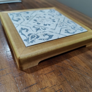 May include: Square wooden coaster with a decorative tile insert. The tile features a grey and white floral pattern. The coaster has a light brown wooden frame and is on a wooden surface.