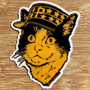 以下が含まれることがあります： 星と文字「P」が付いた黒と金の帽子をかぶった猫のステッカー。猫は黒と金の毛皮と大きな目をしています。ステッカーには白い縁取りがあり、アーティストのサインが入っています。