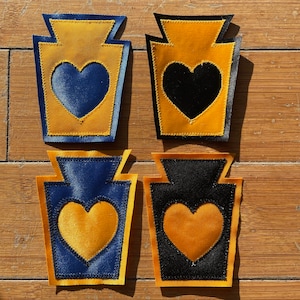 Könnte beinhalten: Vier Stoffaufnäher in Ketchupflaschenform, jeweils mit einem Herzdesign. Die Aufnäher haben eine goldene Flaschenkontur mit einem blauen oder schwarzen Herzen. Der Hintergrund ist eine Holzoberfläche.