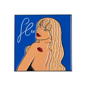 May include: Square enamel pin featuring a stylized illustration of a woman with blonde hair, red lips, and green eyes against a blue background. The pin includes the word "fe" in a cursive font and a red lip design.