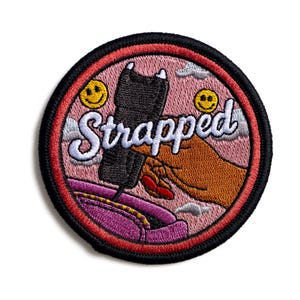 May include: Embroidered patch with the word "Strapped" in white script. The patch features a black object held by a brown hand, a pink background, and a red border. Smiley faces and clouds are also included.