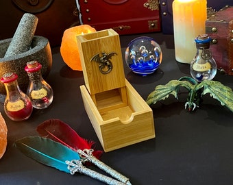 May include: A wooden dice tower with a dragon design on the front. The tower is open and shows the dice rolling tray inside. There are two glass bottles with red liquid and labels that say "Healing" and a glass bottle with blue liquid and a label that says "Mana". There are also two feathers with silver tips and a small green dragon figurine.