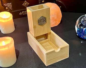May include: A wooden dice tower with a drawer for storing dice. The tower has a metal D20 symbol on the front. The tower is made of light brown wood and has a natural finish.