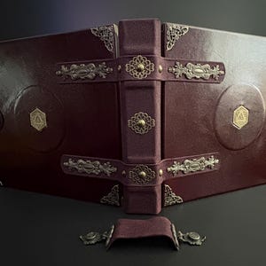 Könnte beinhalten: Ein burgunderfarbenes, ledergebundenes Buch mit verzierten Metallecken und -verschlüssen. Das Buch hat einen zentralen Buchrücken mit einem passenden burgunderfarbenen Riemen und dekorativen Metallverzierungen. Der Einband enthält kreisförmige Designs mit goldfarbenen Emblemen.