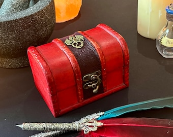 May include: A small red wooden treasure chest with a brass dragon design on the lid and a brass latch. The chest is sitting on a black surface with a quill pen and feathers.
