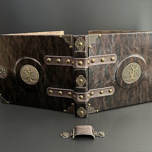 May include: A brown leather-bound book with brass detailing and a tree of life emblem. The book features a decorative clasp and metal corner protectors. The spine is dark brown with ornate metal details. The book is open, revealing the pages.