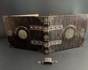 May include: A brown leather-bound book with brass accents and a tree of life emblem. The book has a decorative clasp and metal corner protectors. The spine is dark brown with ornate metal details. The book is open, revealing the pages.