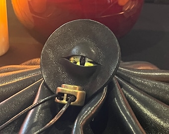 Puede incluir: Una bolsa de cuero negro con una solapa redonda de cuero negro con un diseño de ojo amarillo. La bolsa tiene un cierre de latón y un cierre de cordón.