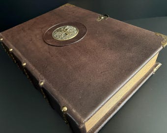 May include: A brown leather-bound journal with a brass emblem and corner protectors. The journal's cover has a textured appearance, and the pages have a golden hue. The journal is closed and appears to be new.
