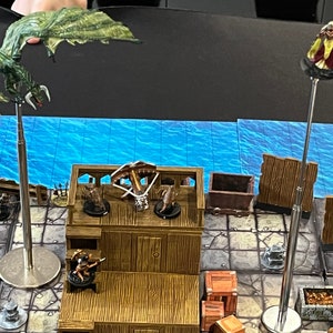 Puede incluir: Una escena de juego de mesa con figuras en miniatura, incluyendo un dragón verde, una figura con una ballesta y una figura con una capa azul. Las figuras están en una plataforma de madera con un fondo de agua azul.