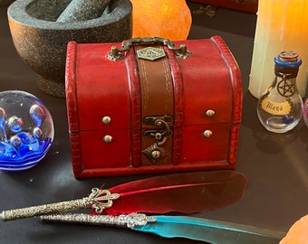 May include: A red wooden treasure chest with a brown leather strap and metal accents. The chest is closed and has a metal latch. There are two quill pens with feathers on a black surface.