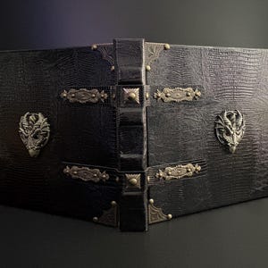 Könnte beinhalten: Ein schwarzes Buch aus Kunstleder mit einem strukturierten Reptilienmuster. Das Buch hat verzierte Metalleckenschützer, dekorative Verschlüsse und Drachenkopfverzierungen. Der Buchrücken ist ebenfalls schwarz und hat ein erhabenes Design.